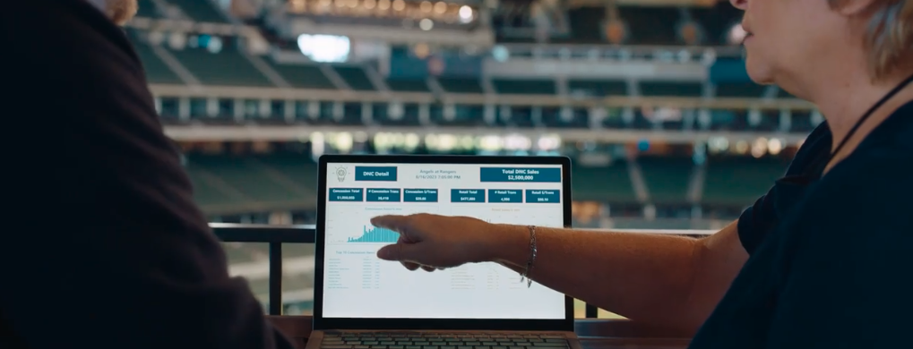 Open laptop showing a Tableau dashboard with a woman pointing at it. The scene is taking place in the seating area of an stadium.