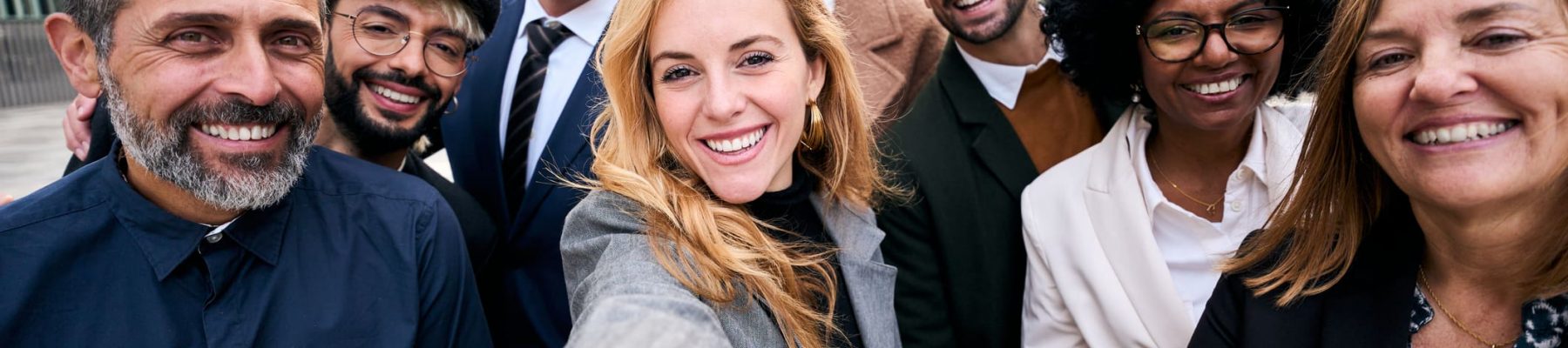 Selfie of a cheerful team of diverse business people in formal suit looking smiling at camera gathered outside the work building. Positive emotion team work celebrating success together